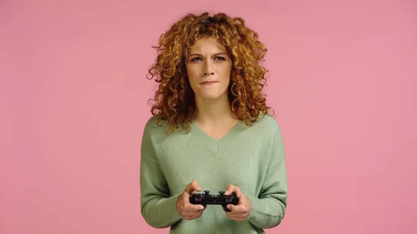 KYIV, UKRAINE - DECEMBER 22, 2021: tense redhead woman playing video game with joystick isolated on pink — Stockfoto