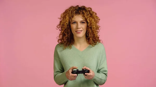 KYIV, UKRAINE - DECEMBER 22, 2021: smiling redhead woman gaming with joystick isolated on pink — Foto stock