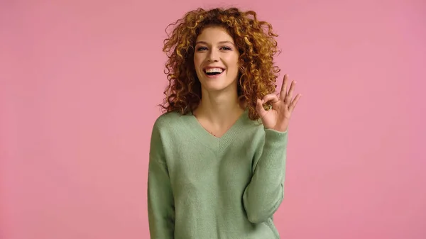Excited woman with red wavy hair showing okay sign while looking at camera isolated on pink — Foto stock
