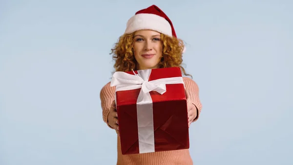 Розмита усміхнена жінка в капелюсі Санта, що показує велику подарункову коробку ізольовано на синьому — стокове фото