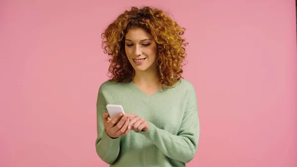 Joyful woman with curly and red hair using mobile phone isolated on pink — Stock Photo