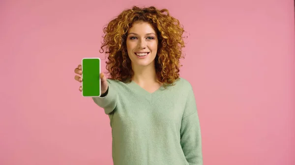 Gai rousse femme regardant caméra tout en montrant smartphone avec écran vert isolé sur rose — Photo de stock