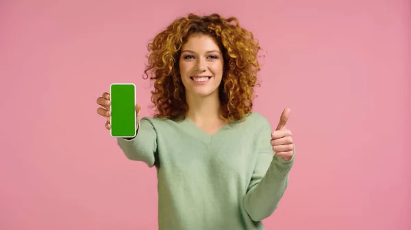 Joyful woman holding cellphone with green screen and showing thumb up isolated on pink — Stockfoto