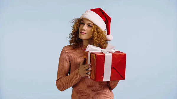 Curious and intrigued woman in santa hat holding large gift box isolated on blue — Stockfoto