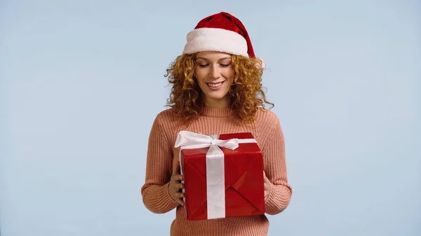 Щаслива руда жінка в капелюсі Санта дивиться на новий рік подарунок ізольовано на синьому — стокове фото