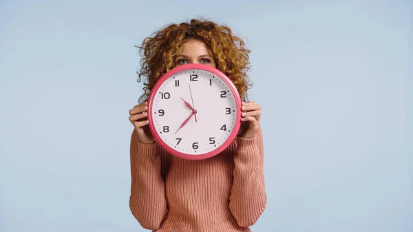 Junge Frau verdeckt Gesicht mit rund um die Uhr, während sie isoliert auf blauem Grund in die Kamera schaut — Stockfoto