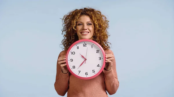 Joyeuse rousse femme tenant horloge ronde blanche tout en regardant la caméra isolée sur bleu — Photo de stock
