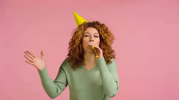 Redhead woman in party cap blowing in party horn and waving hand isolated on pink — Stockfoto