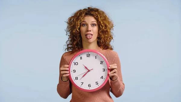 Mujer joven con el pelo ondulado rojo que sobresale de la lengua mientras sostiene el reloj redondo aislado en azul - foto de stock