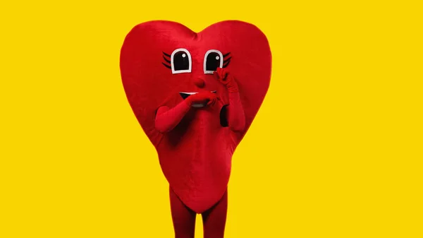 Persona en traje de corazón rojo tocando los ojos de dibujos animados mientras imita el llanto aislado en amarillo - foto de stock