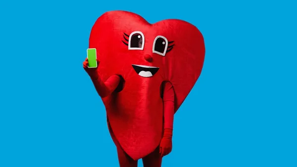 Людина в позитивному костюмі серця тримає смартфон із зеленим екраном ізольовано на синьому — стокове фото