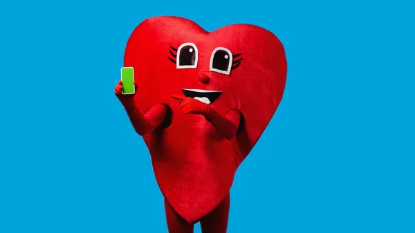 Persona in costume cuore positivo che punta a smartphone con schermo verde isolato su blu — Foto stock