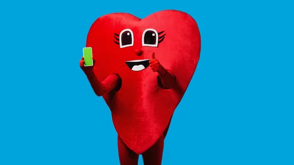 Persona in costume cuore positivo che tiene smartphone con schermo verde e mostra come isolato sul blu — Foto stock