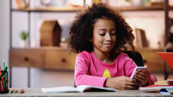 Smiling african american child using smartphone while doing homework — Stock Photo