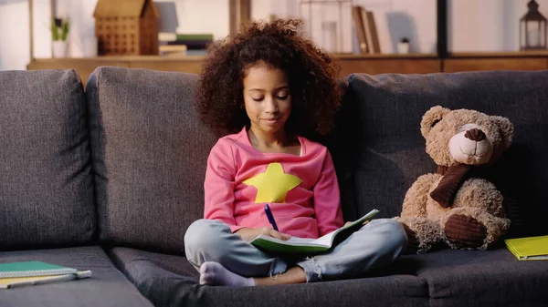 Rizado africano americano niño haciendo tarea en sofá - foto de stock