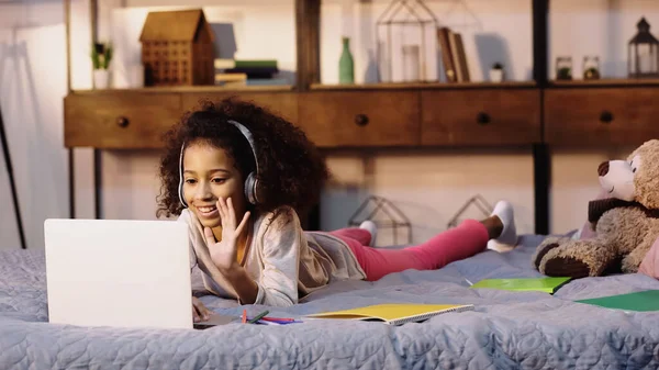 Glückliches afrikanisch-amerikanisches Kind mit Kopfhörer, das Videoanruf auf Laptop im Bett hat — Stockfoto
