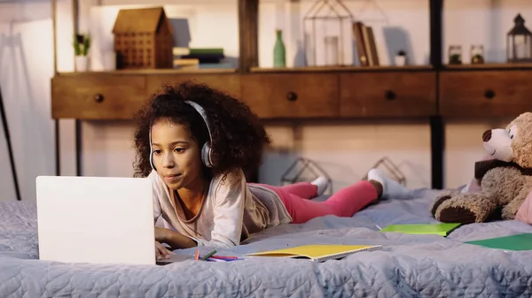 Curly african american child in wireless headphones using laptop in bed — Foto stock