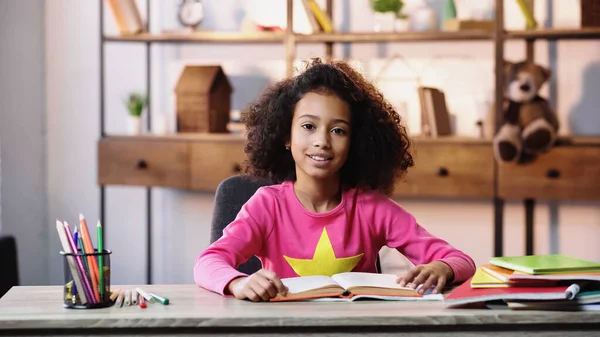 Smiling african american child reading book and looking at camera — Stock Photo