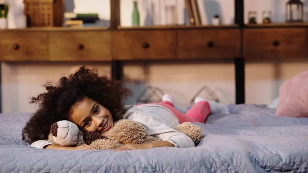 Feliz criança afro-americana deitada com ursinho de pelúcia na cama — Fotografia de Stock