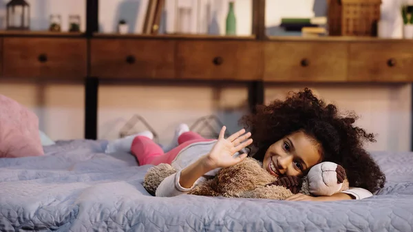 Felice afro americano bambino agitando mano mentre sdraiato con orsacchiotto a letto — Foto stock