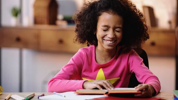 Menina afro-americana feliz lendo livro e rindo em casa — Fotografia de Stock