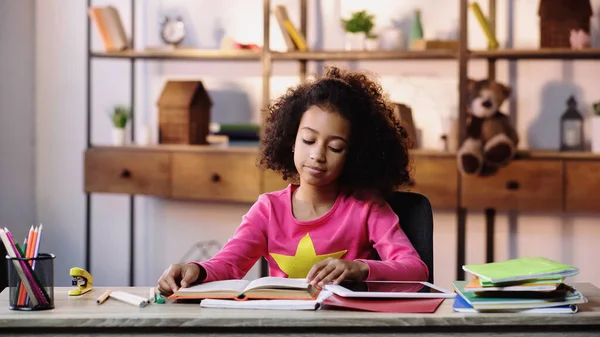 Афроамериканська дівчина читає книгу біля копій і цифрового планшета на столі — стокове фото