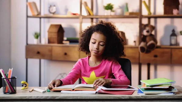 African american girl reading book near copybooks and digital tablet on desk — Stockfoto