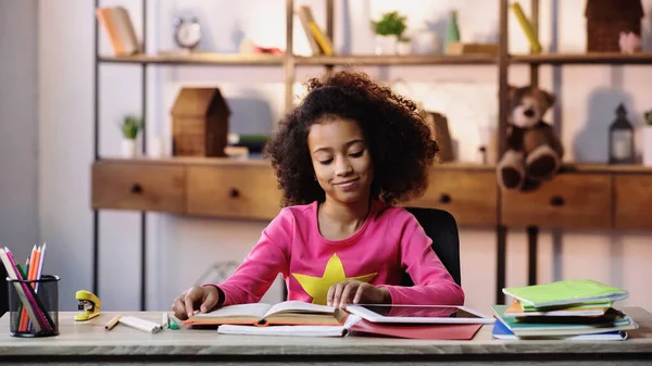 Menina africana feliz livro de leitura perto de cadernos e tablet digital na mesa — Fotografia de Stock