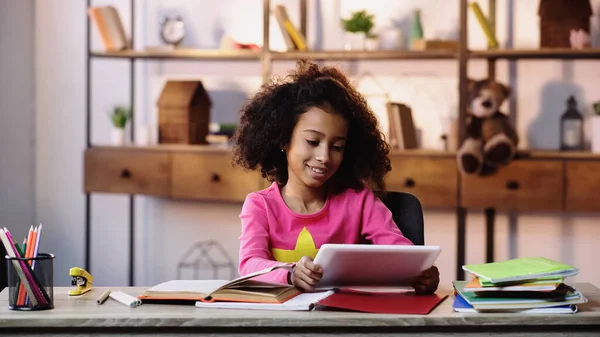 Happy african american girl using digital tablet near notebooks on desk — Stock Photo