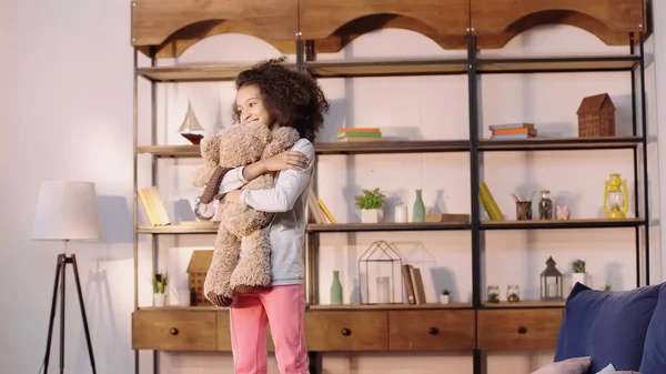 Cheerful african american girl hugging teddy bear in living room — Fotografia de Stock