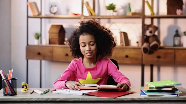Feliz menina afro-americana ler livro perto de cadernos na mesa — Fotografia de Stock