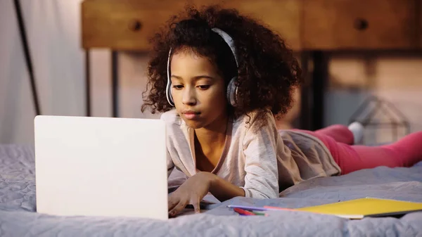 Niño afroamericano rizado en auriculares inalámbricos usando computadora portátil en la cama - foto de stock