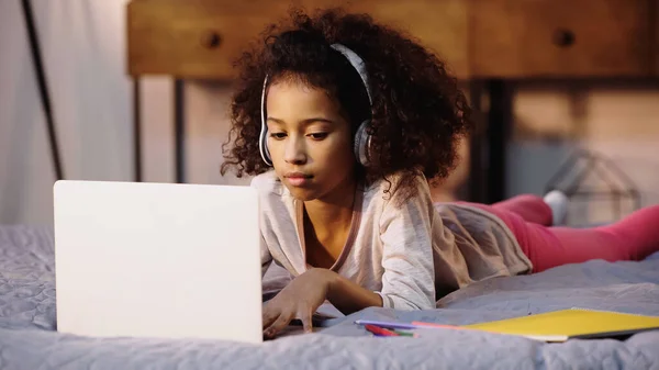 Curly african american child in wireless headphones using laptop in bed — Stock Photo