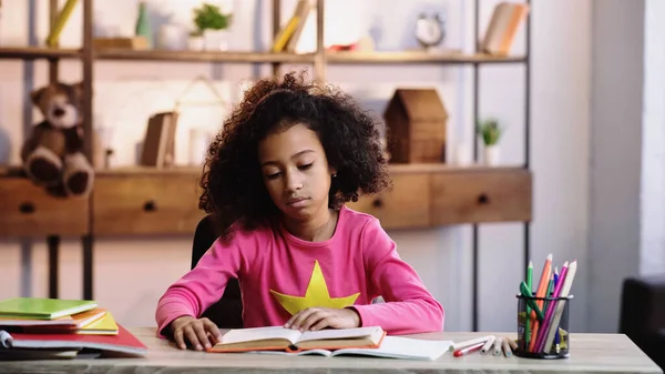 Curly african american schoolkid reading book at home — Stock Photo