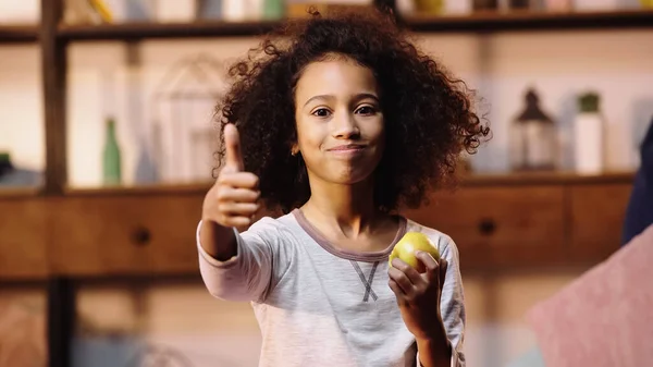 Niño afroamericano mostrando el pulgar hacia arriba mientras come manzana - foto de stock