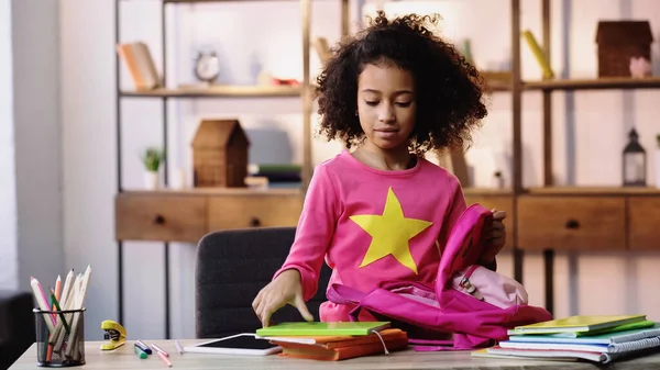 Curly african american child packing backpack near digital tablet on table — Stockfoto