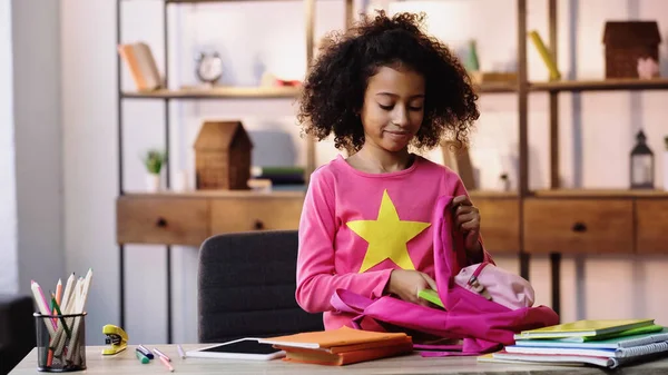 Curly african american child putting notebooks in backpack — Stockfoto