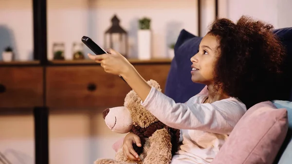 Glücklich afrikanisch-amerikanisches Kind wechselt Sender, während es mit Teddybär fernsieht — Stockfoto