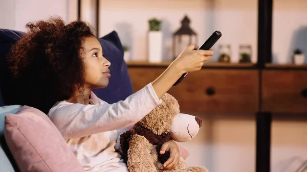 African american child switching channels while watching tv with teddy bear — Stock Photo