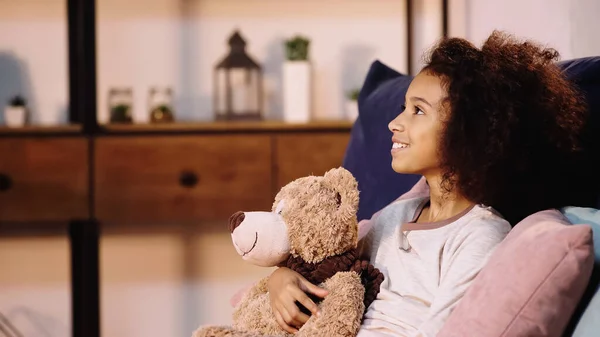 Vista lateral de un niño afroamericano feliz sosteniendo un osito de peluche y mirando hacia otro lado - foto de stock