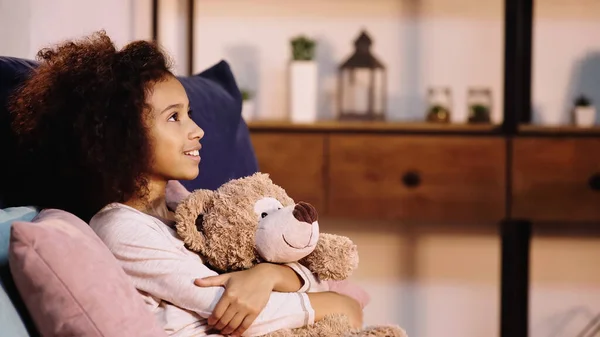 Side view of cheerful african american child holding teddy bear and looking away — Foto stock