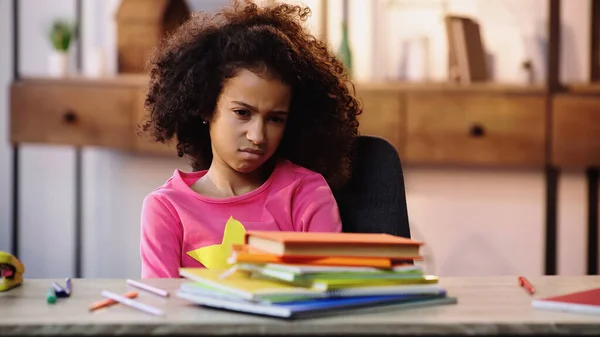 Displeased african american schoolchild looking at notebooks while doing homework — Stock Photo