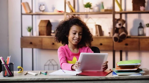 Smiling african american girl using digital tablet near notebooks on desk — Stock Photo