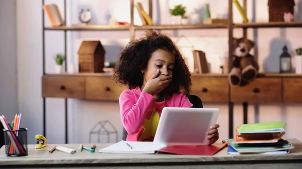 Кучерява афроамериканська дівчина використовує цифровий планшет, прикриваючи рот біля блокнотів на столі — стокове фото