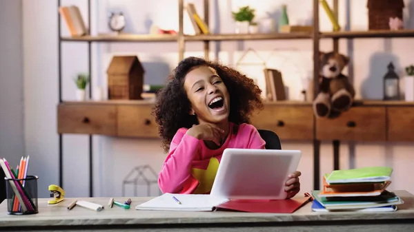 Menina americana africana alegre usando tablet digital e rindo perto de cadernos na mesa — Fotografia de Stock
