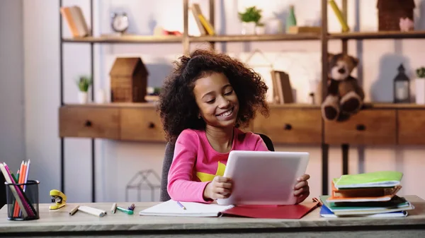 Menina americana africana alegre usando tablet digital perto de cadernos na mesa — Fotografia de Stock