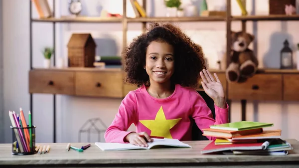 Heureux afro-américaine fille agitant la main et regardant la caméra tout en faisant des devoirs — Photo de stock