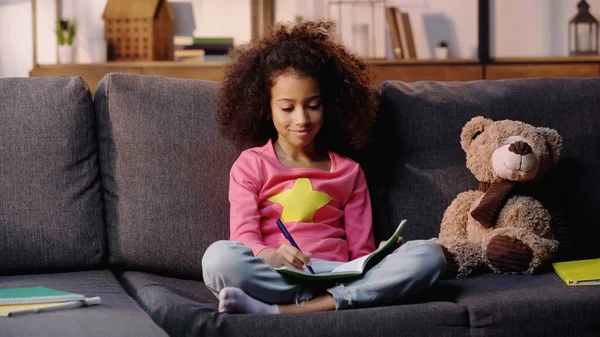 Curly african american child writing in notebook near teddy bear on sofa — Stock Photo