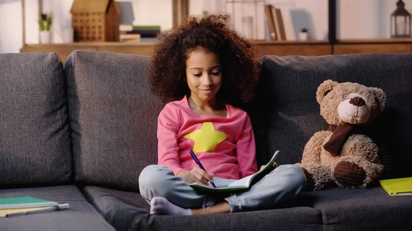 Niño afroamericano rizado escribiendo en cuaderno cerca de oso de peluche en sofá - foto de stock