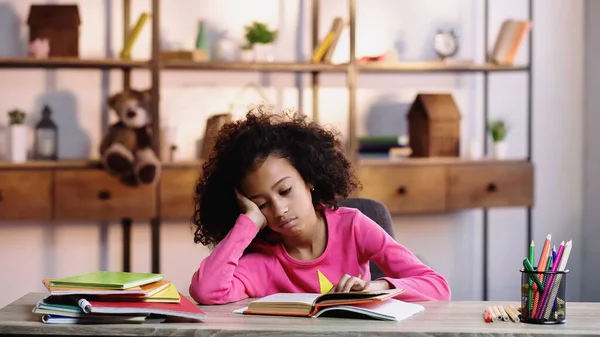 Unzufriedenes afrikanisch-amerikanisches Kind schaut sich Buch an, während es Hausaufgaben macht — Stockfoto